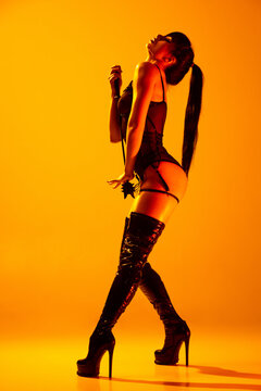 Studio shot of sexy young woman wearing leather lingerie posing isolated on yellow background in neon light. Concept of beauty, love