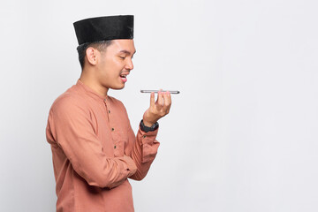Smiling young Asian Muslim man using the voice assistant on mobile phone isolated over white background