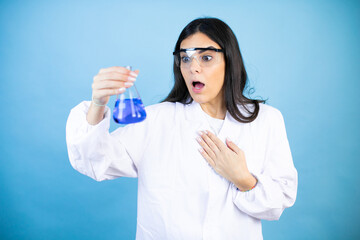 Young brunette woman wearing scientist uniform holding test tube over isolated blue background afraid and shocked with surprise and amazed expression, fear and excited face.