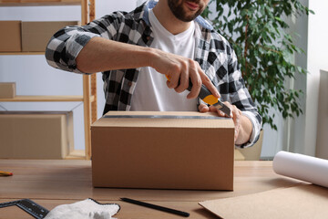 Man using utility knife to open parcel at wooden table indoors, closeup