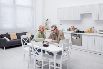 Senior woman talking to husband holding paper near coffee and laptop in kitchen.