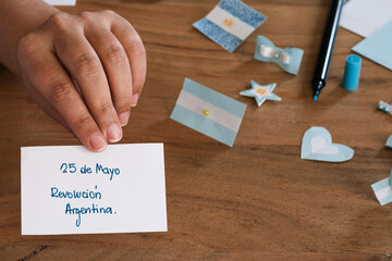 hand holding a piece of paper written in Spanish 25 de mayo revolucion Argentina, in the background colors of light blue and white