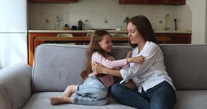 Young attractive mom and little daughter tickling each other seated on sofa at home, play together spend weekend carefree leisure have fun looking happy and overjoyed. Motherhood, family bond concept