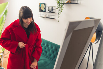 Caucasian brown-haired woman trying out warm red coat in front of mirror. Online shopping concept. High quality photo