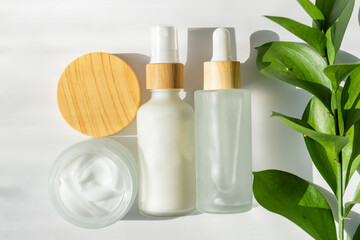 White cosmetic bottles and open jar with face cream on white background with green ruscus leaves. Skin care, body treatment, beauty concept. Unbranded packaging for skincare treatment