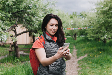 Joyful female tourist with backpack using map during travel in recreational park area, happy hipster girl connecting to roaming internet for making online travel video vlog