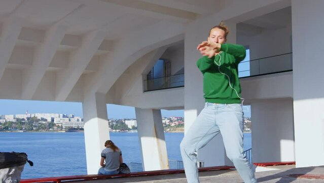 Girl in jeans listening music in earphones and performing modern dances on embankment under roof. Female dancer shows dancing movements of hip-hop, contemporary, shuffle, body wave at seafront stage