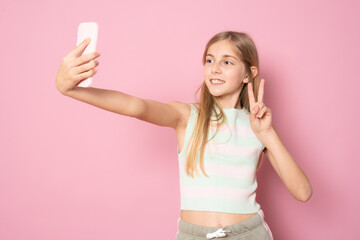 Excited little blonde kid girl 11-12 years old in summer clothing doing selfie isolated on pink...