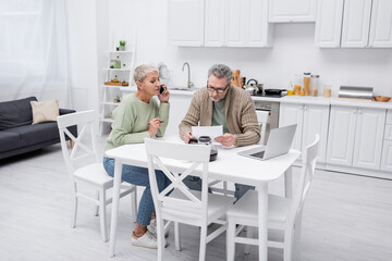 Senior woman talking on smartphone while husband holding paper in kitchen.