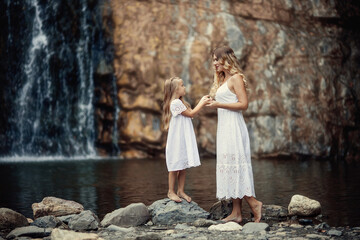 Beautiful mother and daughter wearing white dresses in the mountains near the river on the background of a waterfall.