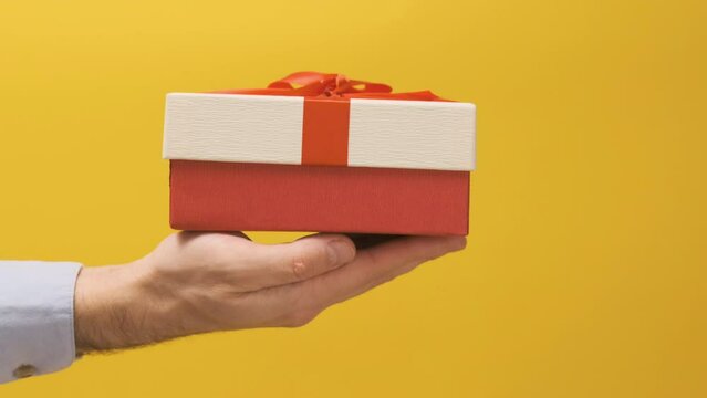 Mans hand holds red gift box in palm of his hand and lowers it down the screen on bright yellow background. Giving gift or surprise.
