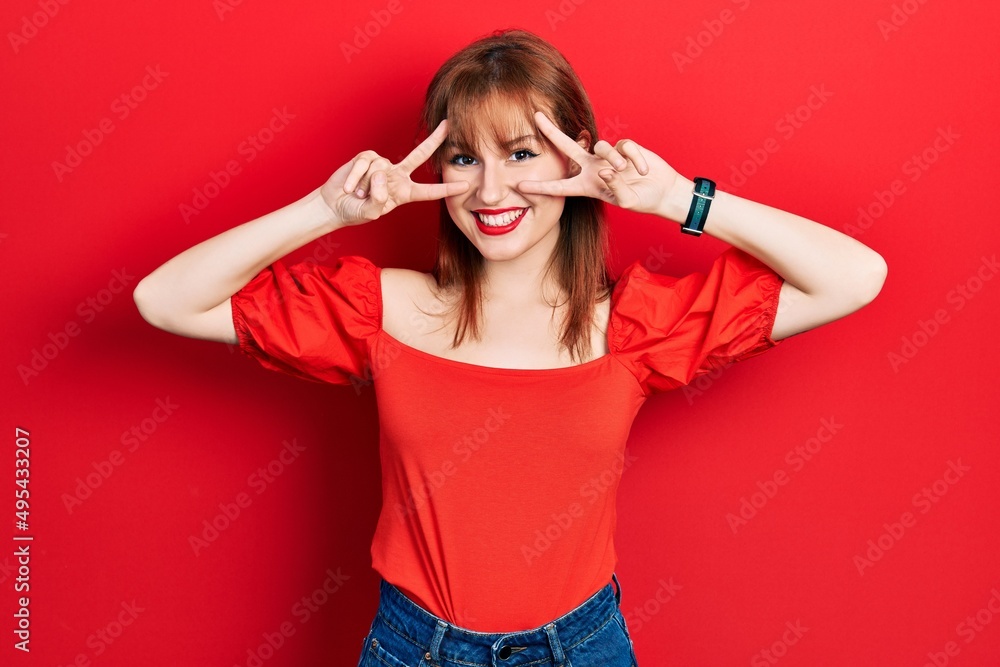 Sticker redhead young woman wearing casual red t shirt doing peace symbol with fingers over face, smiling ch