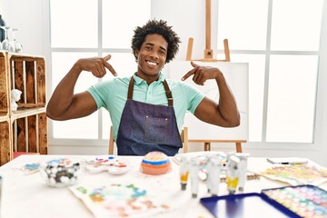 Young african american man sitting on the table at art studio looking confident with smile on face, pointing oneself with fingers proud and happy.