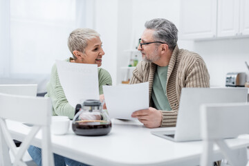 Pensioners holding documents near devices and coffee in kitchen.