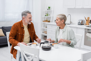 Cheerful mature man talking to wife near tasty pancakes and coffee in kitchen.