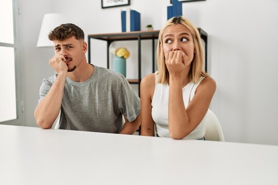 Young beautiful couple wearing casual clothes sitting on the table at home looking stressed and nervous with hands on mouth biting nails. anxiety problem.
