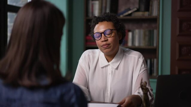 Young woman lawyer talking to client and sitting at table in company spbas. 4k Close view of american african american businesswoman talks to client and looks with smile, reads lawsuit or legal case
