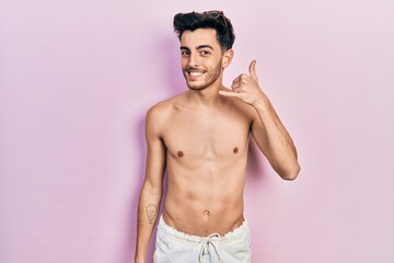 Fototapeta premium Young hispanic man wearing swimwear shirtless smiling doing phone gesture with hand and fingers like talking on the telephone. communicating concepts.
