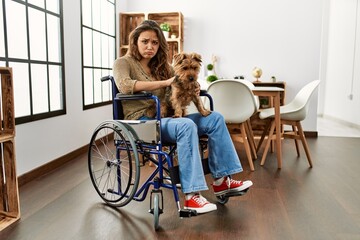 Young hispanic girl sitting on wheelchair at home skeptic and nervous, frowning upset because of...