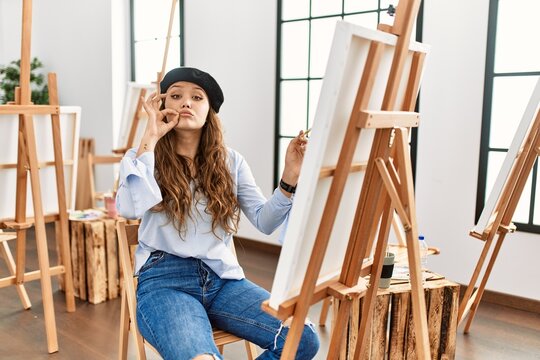 Young hispanic artist woman painting on canvas at art studio mouth and lips shut as zip with fingers. secret and silent, taboo talking