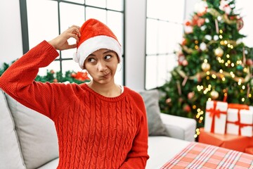 Young hispanic woman with short hair wearing christmas hat sitting on the sofa confuse and...