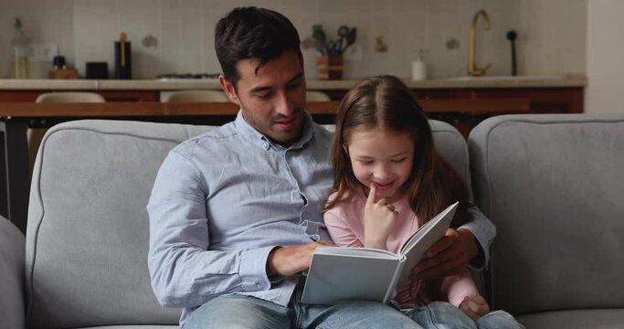 Young dad and his preschool 6s daughter relaxing on sofa read book looking at pictures spend leisure enjoy hobby and pastime. Fatherhood, upbringing, kids development and intellectual growth concept