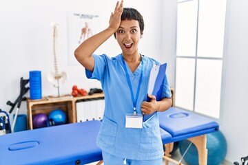 Young hispanic woman with short hair working at pain recovery clinic surprised with hand on head...