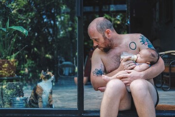 Father feeding his baby with a bottle while cat looking