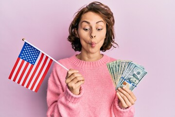 Young brunette woman holding united states flag and dollars making fish face with mouth and...