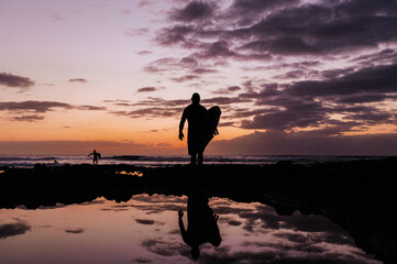Unrecognizable surfers get out of the water during sunset or sunrise. Reflection of dramatic sky.
