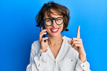 Young brunette woman having conversation talking on the smartphone smiling with an idea or question pointing finger with happy face, number one