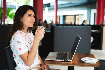 Side view smiling woman with positive expression holding a cup of coffee looking at distance. Female sitting at cafe table with computer laptop.