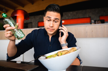 Portrait confused, uncertain male talking at phone sitting at table while have healthy lunch with salad.