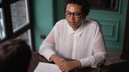 Two young women talking and smiling while sitting at table in modern office spbas. 4k Portrait of american african female talks to client and looks with smile, works and sits at desk with lawsuit and - Powered by Adobe
