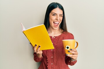 Young hispanic woman reading book drinking cup of tea winking looking at the camera with sexy...