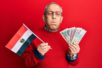 Handsome senior man with grey hair holding egypt flag and egyptian pounds banknotes puffing cheeks with funny face. mouth inflated with air, catching air. - obrazy, fototapety, plakaty