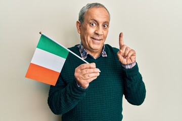Handsome senior man with grey hair holding ireland flag smiling with an idea or question pointing finger with happy face, number one