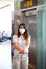 Vertical shot young caucasian woman in casual clothes and face mask standing looking at camera. Female with trolley in the airport or station. Travel during pandemic concept.