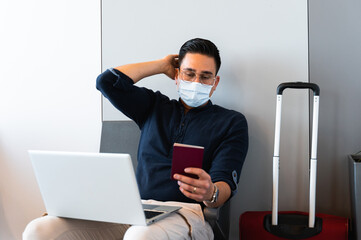Young man with face mask sitting in waiting lounge at airport. Businessperson using computer puts hand behind head looking at the passport with worried face. Travel and documentation problems concept.
