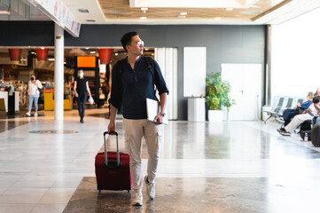 Front view handsome hispanic man with trolley suitcase walking at the airport. Travel concept.