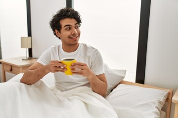 Fototapeta na wymiar Young hispanic man drinking coffee sitting on the bed at bedroom.
