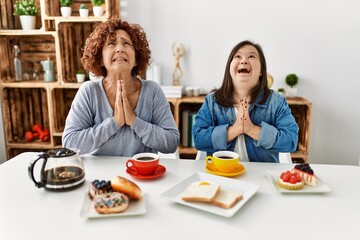 Family of mother and down syndrome daughter sitting at home eating breakfast begging and praying with hands together with hope expression on face very emotional and worried. begging.