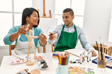 Young latin painter couple smiling happy using manikin sitting on the table at art studio