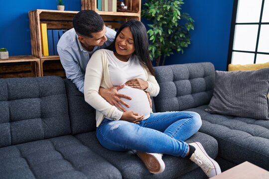 Young latin couple expecting baby hugging each other sitting on sofa at home