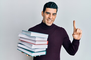 Handsome hispanic man holding a pile of books smiling with an idea or question pointing finger with happy face, number one