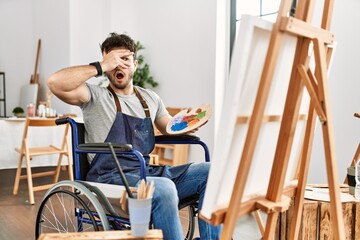 Young hispanic man sitting on wheelchair painting at art studio peeking in shock covering face and eyes with hand, looking through fingers with embarrassed expression.