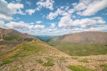 Scenic view from mountain ridge to green forest valley among mountain ranges and hills on horizon at changeable weather. Green landscape with sunlit mountain vastness under cumulus clouds in blue sky.