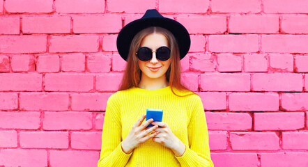 Portrait of young woman with smartphone wearing black round hat, yellow sweater on pink background