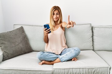 Blonde beautiful young woman sitting on the sofa at home using smartphone pointing with finger up...