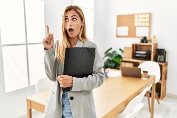 Blonde business woman at the office amazed and surprised looking up and pointing with fingers and raised arms.